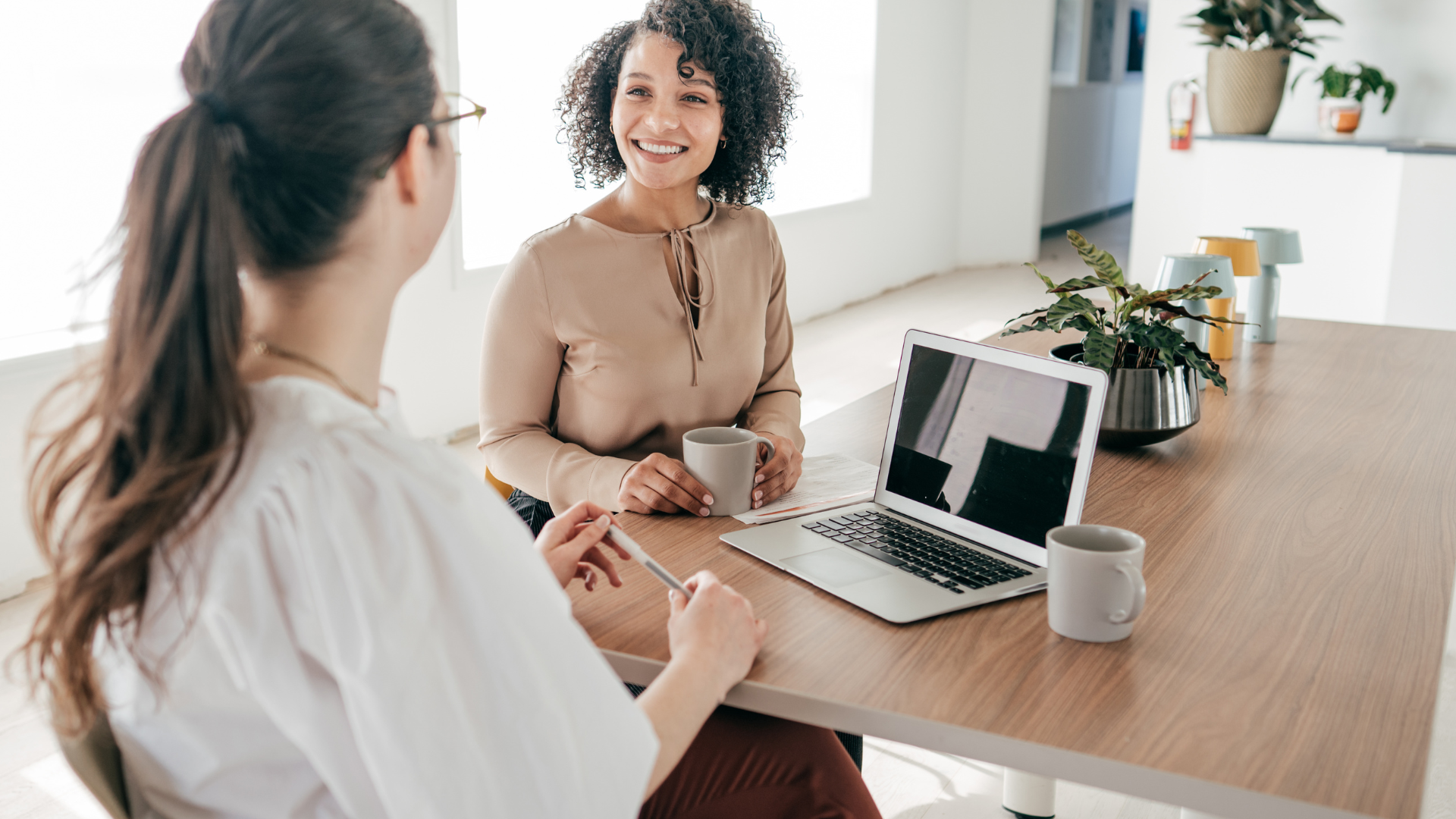 Two women sitting across from each other at work table discussing with coffee and laptop