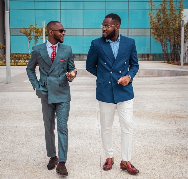 Two black men in business suits top to chat in front of a blue building