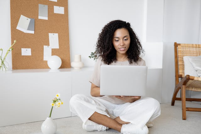 Black woman sitting cross-legged while typing on a computer, and a cork board with notes is behind her.