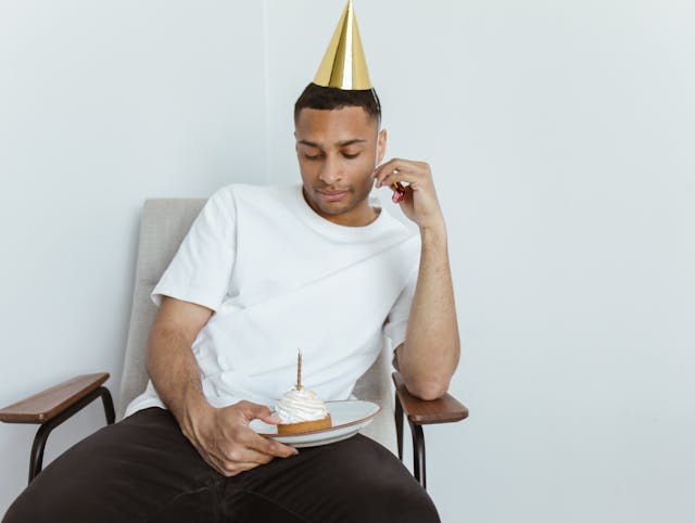 Unhappy man gazes at birthday cupcake while wearing a gold birthday cone hat